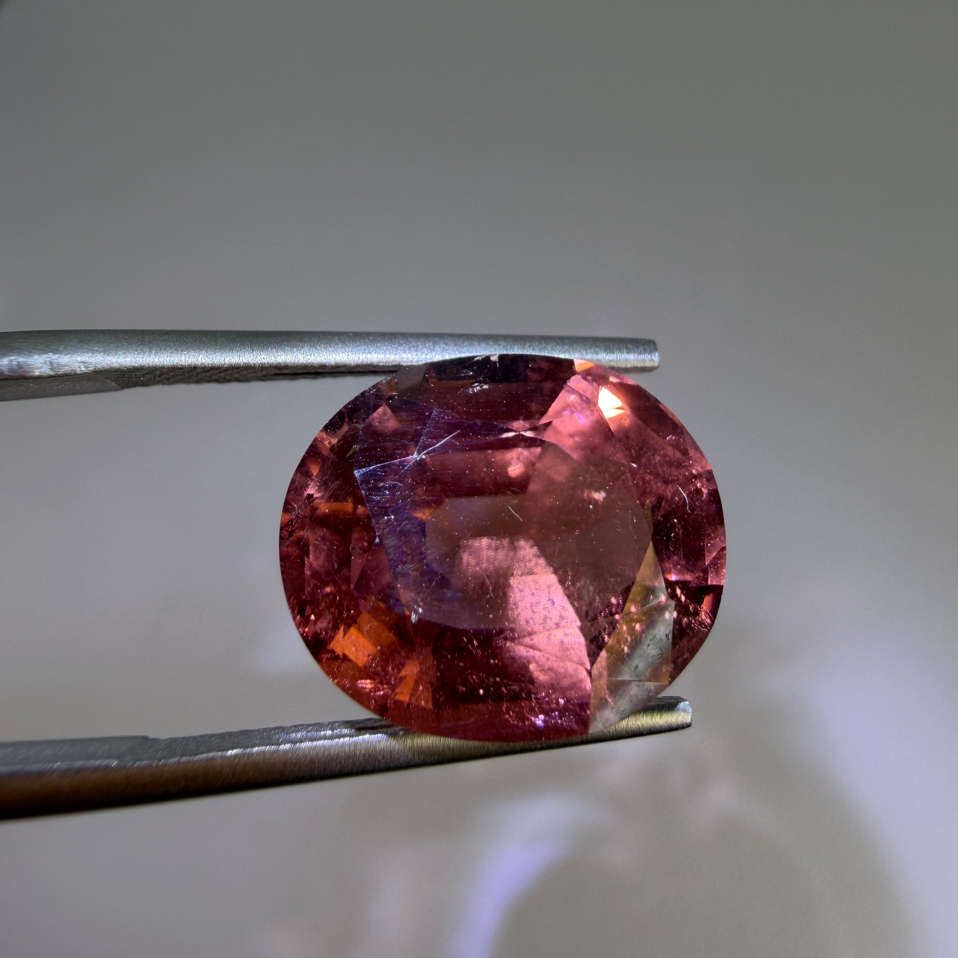 Oval-shaped pinkish-red gemstone held between tweezers against a neutral background